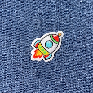 May include: A colorful embroidered iron-on patch of a cartoon rocket ship with a white body, red and yellow flames, and blue and green details.