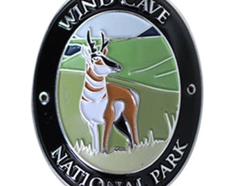 May include: A black and silver oval-shaped metal pin with the words "Wind Cave National Park" around the edge. The center of the pin features a colorful illustration of a pronghorn antelope standing in a grassy field.