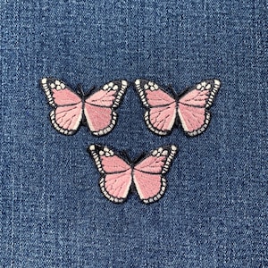 May include: Three embroidered butterfly patches with pink wings and white and black detailing are arranged on a blue denim background. The butterflies have a delicate, detailed design, perfect for adding to clothing or accessories.