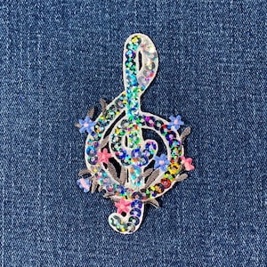May include: A decorative sequined treble clef patch with a floral design. The musical note is adorned with iridescent sequins and surrounded by small blue, pink, and yellow flowers. The patch is set against a blue denim background.