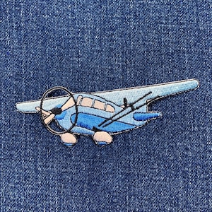 May include: Embroidered patch of a light blue airplane with a dark blue propeller and black accents. The patch is on a blue denim background. The airplane design features a propeller, windows, and a tail.