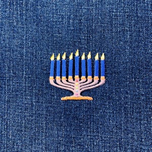 May include: Embroidered menorah patch featuring a pink base with blue candles and yellow flames. The patch is set against a blue denim background. This patch is a decorative item.