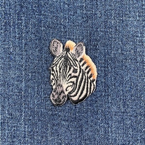 May include: Embroidered zebra head patch with black and white stripes, tan mane, and gray ears. The patch is on a blue denim background. This patch can be sewn or ironed on to clothing or accessories.