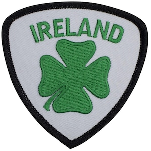 May include: A triangular embroidered patch with a white background and a thick black border. The patch features the word IRELAND in green capital letters arched above a green four-leaf clover centered on the white fabric surface.