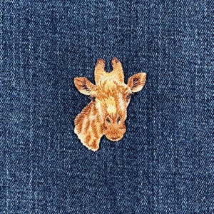 May include: Embroidered patch featuring a giraffe head with detailed brown and tan fur patterns. The patch is set against a dark blue denim background, showcasing the texture of the fabric. This patch is suitable for clothing or accessories.