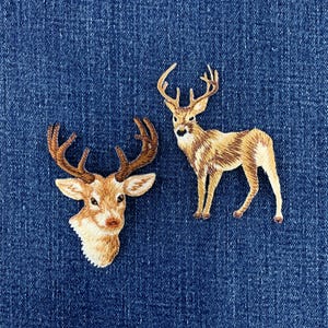 May include: Two embroidered patches featuring deer designs. One patch shows a detailed deer head with brown antlers, while the other depicts a full-body standing deer with large antlers. Both patches are rendered in shades of tan and brown.
