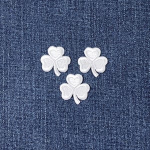 May include: Three white embroidered shamrock patches on a blue denim background. Each shamrock has three heart-shaped leaves and a small stem. The patches are evenly spaced and slightly angled.