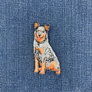 May include: An embroidered patch featuring a blue heeler dog with a mottled grey and white coat, tan legs, and a tan face. The dog is depicted in a sitting position with its mouth slightly open, showing a pink tongue, and pointed ears.