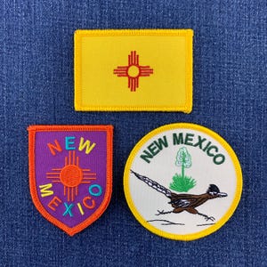 以下が含まれることがあります： ニューメキシコ州をテーマにした3つの刺繍ワッペン。赤いジアのシンボルが描かれた黄色い長方形、赤いジアのシンボルと「NEW MEXICO」の文字が入った紫色の盾型、そしてロードランナーとユッカが描かれた白い円形のワッペン。