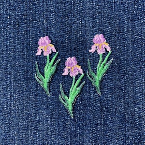May include: Three embroidered patches featuring purple iris flowers with yellow centers and green stems and leaves. The patches are arranged in a triangular formation against a dark blue denim fabric background with a visible woven texture.
