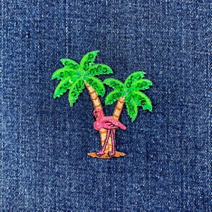 May include: An embroidered patch featuring a pink flamingo standing between two palm trees. The palm trees have green leaves and brown trunks. The patch is set against a blue denim background.