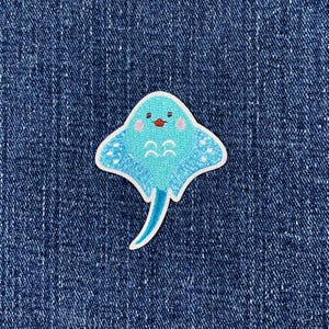 May include: A blue stingray patch with a white border. The stingray has a light blue body with white dots and a darker blue tail. The stingray has a small red mouth and pink cheeks. The patch is on a blue denim background.