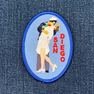 May include: A blue oval embroidered patch with a white background. The patch features a couple kissing, a sailor in a white uniform kissing a woman in a white dress. The text "SAN DIEGO" is written vertically in red on the right side of the patch.