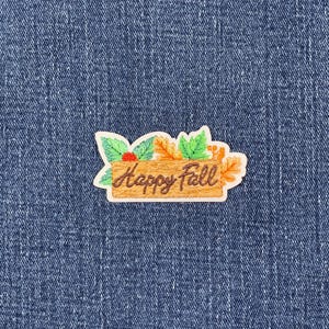 May include: A colourful patch with the words "Happy Fall" in a brown wooden sign design. The patch is decorated with green leaves, orange and yellow leaves, and red berries. The patch is on a blue denim background.