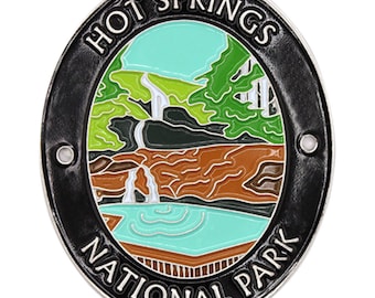 May include: A black and silver metal enamel pin featuring a colorful illustration of Hot Springs National Park. The pin depicts a waterfall flowing into a pool of water with green trees and a blue sky in the background. The text "HOT SPRINGS NATIONAL PARK" is written in a black circle around the illustration.
