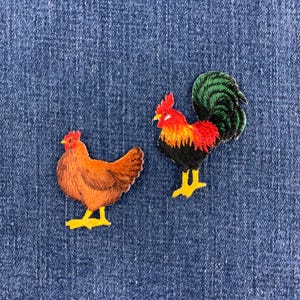 以下が含まれることがあります： 家禽のデザインが施された2つの刺繍ワッペン。1つは黄色い足をした茶色の雌鶏、もう1つは赤い頭、オレンジと黒の体、緑の尾羽、黄色い足をした雄鶏で、どちらも青いデニム生地の上に配置されています。