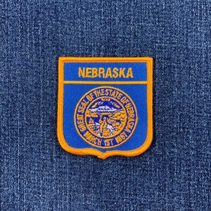 May include: A blue and orange patch featuring the word "NEBRASKA" at the top. The patch has a shield shape with the state seal in the center, surrounded by text. The patch is set against a denim background.