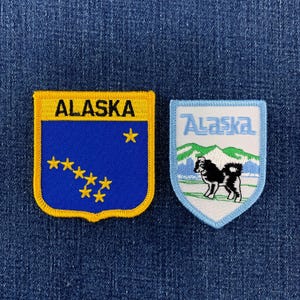May include: Two embroidered patches. The left patch is shield-shaped with a yellow border, blue background, yellow stars, and the word ALASKA in black. The right patch is shield-shaped with a light blue border, white background, green mountains, and a black and white dog.