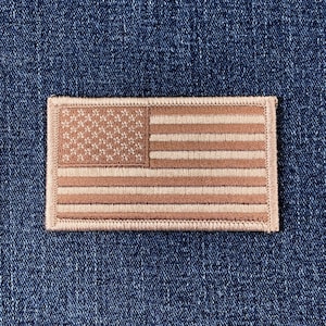 May include: A rectangular embroidered patch depicting the American flag. The design uses tan and off-white thread to create the stars and stripes. The patch is set against a blue denim background, commonly used for clothing.