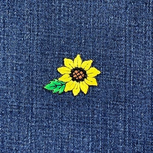 May include: A yellow sunflower patch with a brown center and green leaf is centered on a blue denim background. The patch is embroidered with detailed stitching, creating a textured appearance.