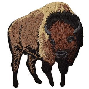 May include: An embroidered patch featuring a standing American bison. The design uses shades of brown, tan, and black thread to depict the animal's thick fur, mane, and sturdy legs. The bison is shown from a front-facing angle with a detailed texture.