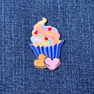 May include: A colorful cupcake patch with a blue liner, topped with swirled frosting in shades of pink, yellow, and blue, and adorned with red heart sprinkles. Two heart-shaped patches, one pink and one gold, are below the cupcake.