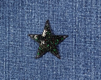 May include: A black sequined star patch with green and gold detailing is set against a blue denim backdrop. The five-pointed star has a textured look, enhancing its visual appeal.
