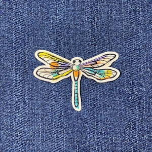 May include: A colourful dragonfly patch with a white border. The dragonfly has wings in shades of yellow, orange, purple, and teal. The body is yellow and orange, and the tail is teal. The patch is on a blue denim background.