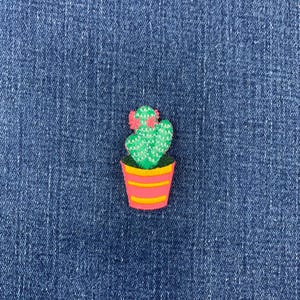 May include: An embroidered patch featuring a green cactus with white cross-stitch detailing and two pink flowers on top. The cactus is planted in a pink pot with horizontal yellow stripes, creating a colorful and detailed decorative accessory.