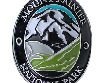 May include: A black and silver oval-shaped metal pin with the words "Mount Rainier National Park" around the edge. The center of the pin features a stylized image of Mount Rainier with a green mountain range in the foreground and a blue sky in the background.