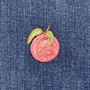 May include: An embroidered peach-shaped patch with a pink and gold glitter design. The patch features green leaves and a brown stem, set against a blue denim background.