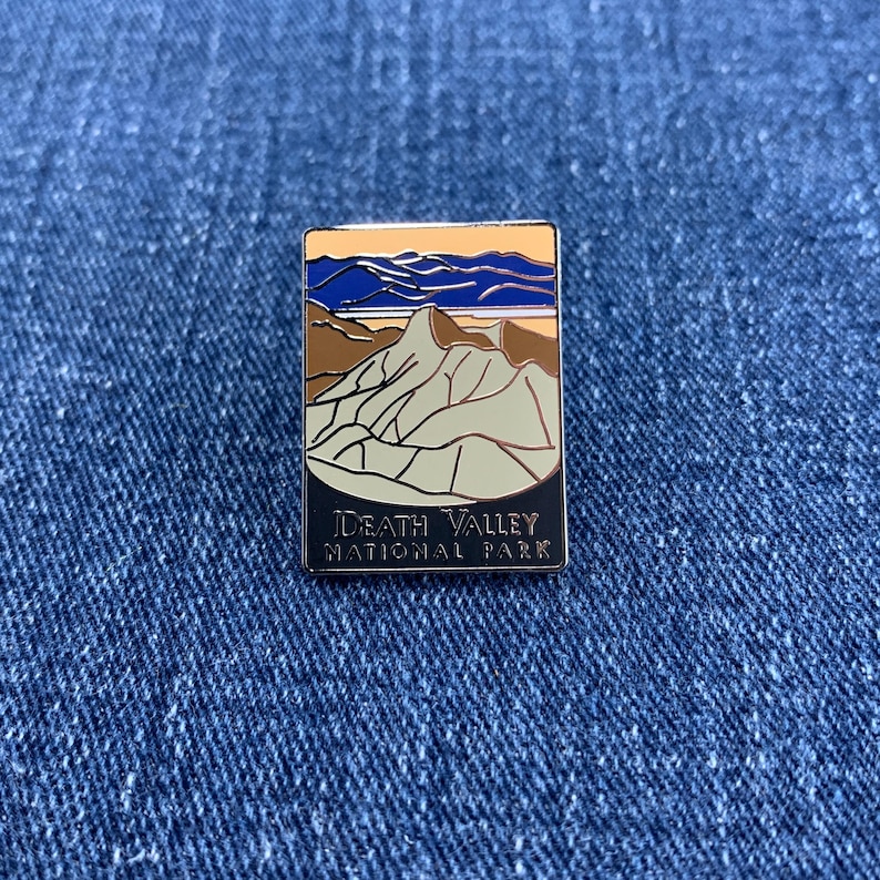 May include: Rectangular enamel pin featuring a landscape design with mountains, sky, and the text "DEATH VALLEY NATIONAL PARK." The pin has a black border and is set against a blue denim background. The colors include beige, brown, blue, and orange.
