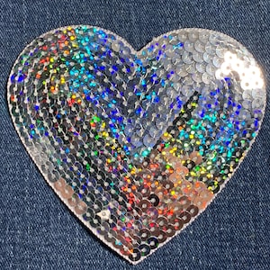 May include: A silver heart-shaped patch covered in iridescent sequins. The sequins are arranged in a pattern that creates a shimmering effect.