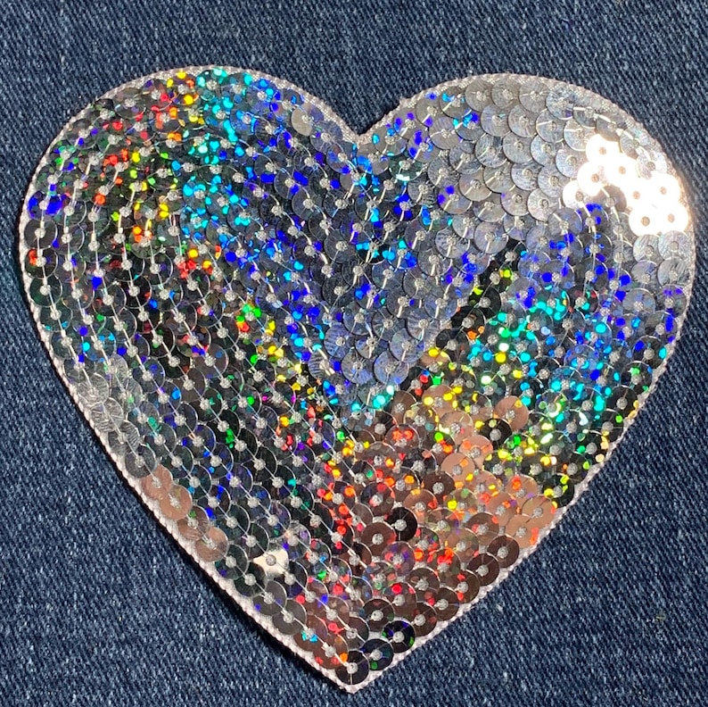 May include: A silver heart-shaped patch covered in iridescent sequins. The sequins are arranged in a pattern that creates a shimmering effect.