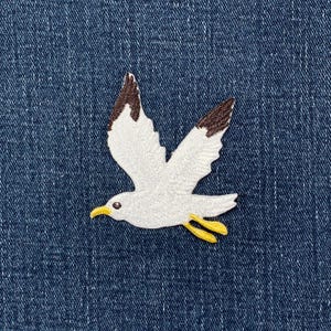 May include: Embroidered patch of a seagull in flight. The bird has white feathers with brown wingtips and yellow feet and beak. The patch is on a blue denim background.