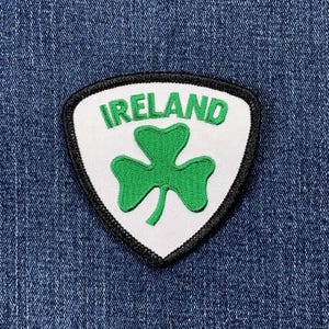 May include: A white, shield-shaped patch with the word "IRELAND" in green letters above a green shamrock. The patch has a black border and is set against a blue denim background.