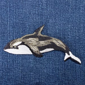 May include: A black and white embroidered patch of an orca whale on a blue denim background.