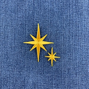 May include: A yellow embroidered patch featuring two eight-pointed stars of different sizes, with a larger star on the left and a smaller star on the right, stitched onto a textured blue denim fabric background with visible diagonal weave lines.