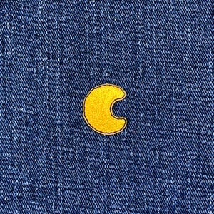 May include: A yellow crescent moon patch with a black border on a blue denim background. The embroidered patch has a textured appearance, adding a decorative element to the fabric. The patch is centered.