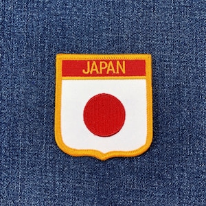 May include: Embroidered patch featuring the Japanese flag design. The patch has a white background with a red circle in the center, a red top border with the word "JAPAN" in gold letters, and a yellow border.