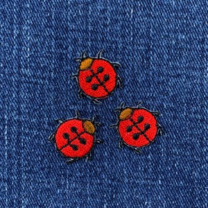 May include: Three red and black embroidered ladybug patches on a blue denim background.