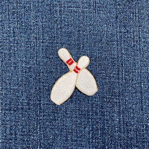 May include: Embroidered patch featuring two white bowling pins with red stripes, set against a blue denim background. The pins are slightly overlapping, suggesting motion or a strike. The patch has a gold border.