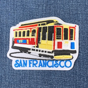 May include: A white embroidered patch with a red, yellow, and black San Francisco cable car. The patch has the text "SAN FRANCISCO" in blue embroidery and "SF" in blue embroidery on the side of the cable car.