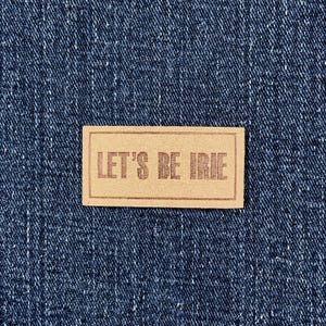 May include: A rectangular, light brown label with the text "LET'S BE IRIE" in a darker brown font. The label is set against a textured, dark blue denim background.