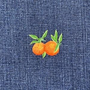 May include: Embroidered patch featuring two orange citrus fruits with green leaves. The patch is sewn onto a blue denim fabric, creating a contrast between the bright colors of the fruit and the dark background.
