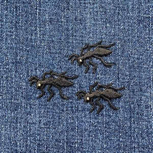 May include: Three black embroidered ant patches with white eye details are arranged on a blue denim background. The patches are detailed with legs, antennae, and body segments. These patches are suitable for clothing or accessories.