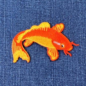 May include: Embroidered patch of a koi carp in orange and yellow hues, set against a blue denim backdrop. The fish is intricately stitched with a small eye and fine details.