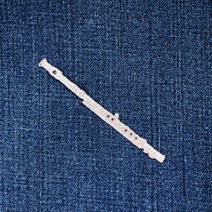 May include: A white, paper-like flute cutout lies diagonally on a textured, dark blue denim background. The flute has several small, dark circles representing the finger holes. The instrument's design is simple and stylized.