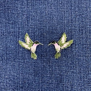May include: Two embroidered hummingbird patches featuring green wings, white underbellies, and pink throat accents. The birds are depicted in flight with wings spread, showcasing detailed stitching and vibrant thread colours against a dark blue denim fabric.