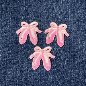 May include: Three pink ballet slipper patches with white accents and rhinestone details are arranged on a blue denim background. Each patch features a bow at the top.